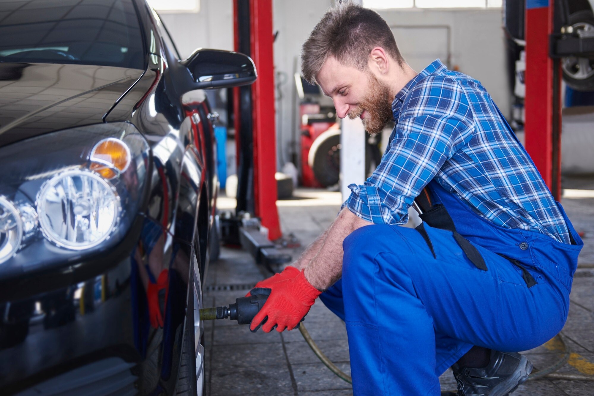 Auto mechanic in blue overalls kneels by a car, using a power tool on the front wheel in a workshop.