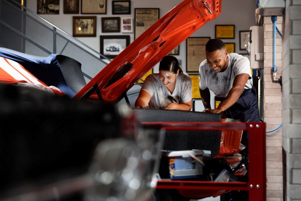 Two people inspect a red sports car with its hood raised in a workshop.