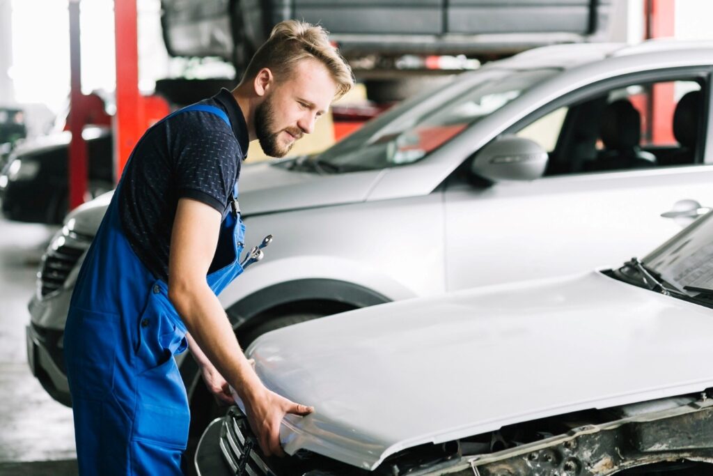 Auto mechanic in blue overalls inspecting the front of a silver car in a workshop